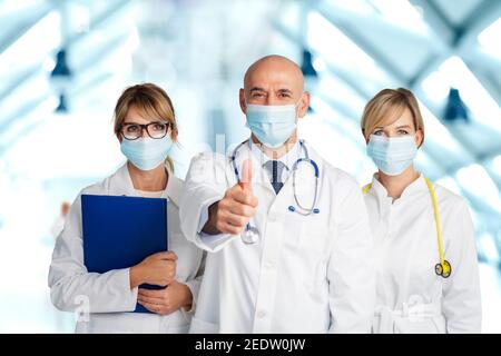 Studioaufnahme des medizinischen Teams, das Gesichtsmasken zur Vorbeugung trägt, und des männlichen Arztes, der Daumen hochzeigt, während er in der Klinik steht. Stockfoto