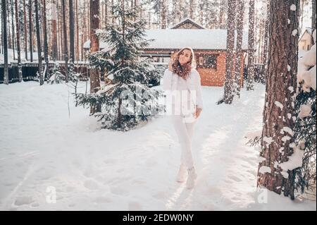 Winter sonnig kalt Porträt im Schneefall der schönen jungen Frau In einer weißen warmen outwear Daunenjacke im Hinterhof Landhaus Holzhaus auf dem Hintergrund der grünen Fichtenkiefer Bäume mit Schnee bedeckt Stockfoto