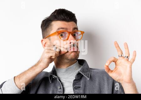 Lustiger junger Mann in einer Brille berührt seinen französischen Schnurrbart und zeigt okay, nicht schlecht Zeichen, genehmigen und wie gutes Produkt, auf weißem Hintergrund stehen Stockfoto