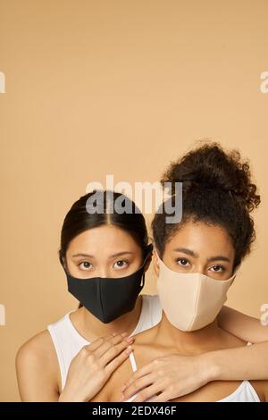 Porträt von zwei liebenswert junge verschiedene Frauen tragen schützende Gesichtsmasken Blick auf Kamera, während zusammen posiert isoliert über beige Hintergrund. Sicherheit, Pandemiekonzept Stockfoto