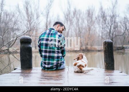 Lächelnder hispanischer Mann mit Flanell und Cavalier King Charles Spaniel Auf einem hölzernen Dock im See Stockfoto