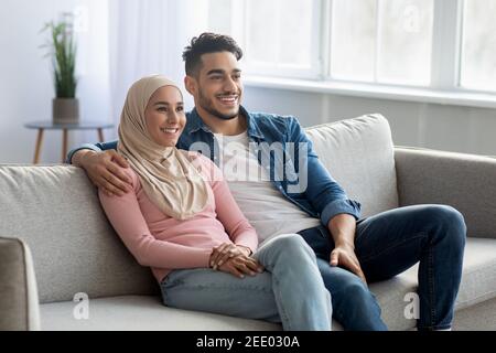 Fröhliches muslimisches Paar, das zu Hause auf der Couch sitzt, Platz kopieren Stockfoto