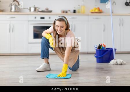 Professioneller Reinigungsservice. Fröhliche junge Dame in Schürze und Gummihandschuhe Waschboden in der modernen Küche Stockfoto