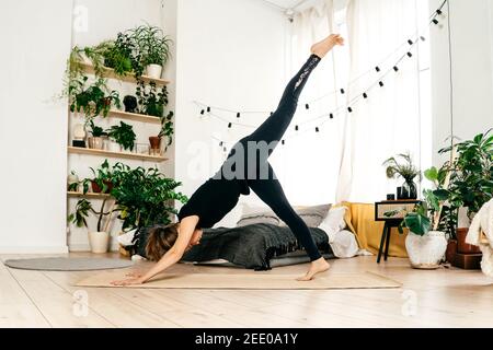 Portrait einer schönen jungen Frau, die drinnen Yoga praktiziert. Frau in schwarzer Sportbekleidung Praxis bei Licht zu Hause. Bett und grüne Heimpflanzen auf dem Hintergrund. Stockfoto