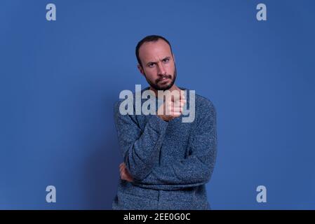Junger Mann mit schweren und wütenden Ausdruck Zeigefinger im Zeichen der Anklage mit Bart und Freizeitkleidung. Im Studio mit blauem Hintergrund Stockfoto