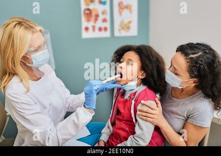 Kind mit ihren Eltern bei Kinderarzttermin. Hausarzt untersucht mit einem Inspektionsspatel den Hals des kleinen Mädchens Stockfoto