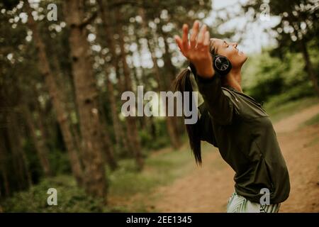 Hübsche junge Frau mit Kopfhörer, die ihre Arme im Wald vorpreading, weil sie draußen trainiert Stockfoto