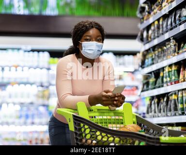 Einkaufen während Coronavirus. Schwarze Frau in Gesichtsmaske Lebensmittel kaufen, überprüfen Lebensmittelliste auf dem Smartphone im Supermarkt Stockfoto