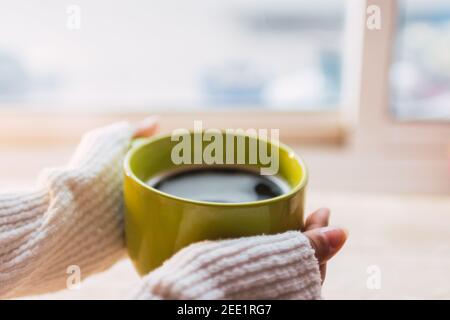 Zwei Hände mit einem cremefarbenen Pullover beim Aufwärmen mit einer grünen Tasse Kaffee aus der Tasse mit einem verschwommenen Hintergrund. Stockfoto