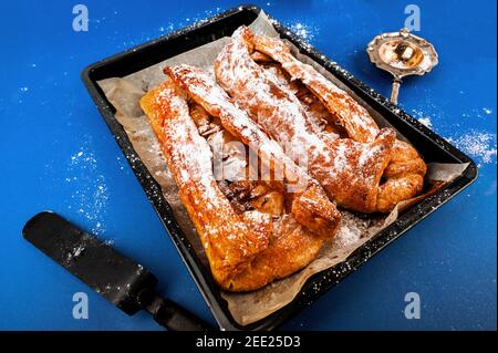 2 Rollen Apfelkuchen "trudel" auf Pfanne, Spachtel und Siebe auf blauem Hintergrund. Stockfoto