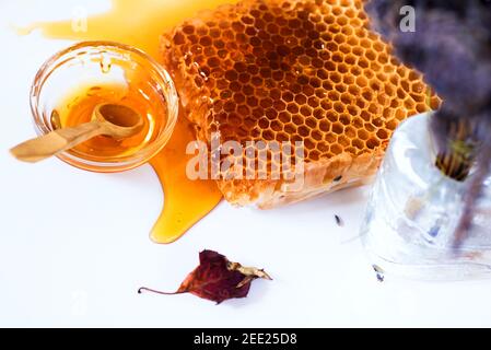 Honigwabe, verschütteter Honig, getrocknetes Blatt, verschwommene Lavendelblüten in Glasvase und kleine Glasschüssel mit Löffel und Honig. Stockfoto