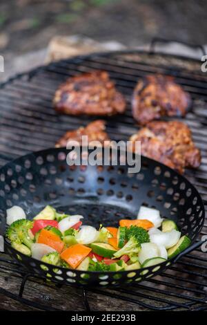 Ein Korb mit Gemüse und marinierten Hähnchenschenkeln kochen auf einem heißen Lagerfeuer. Essen im Freien. Gegrilltes Gemüse. Essen auf einem Campingausflug Stockfoto