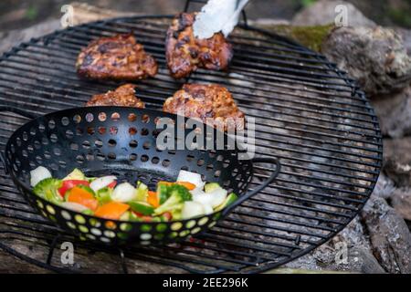 Ein Korb mit Gemüse und marinierten Hähnchenschenkeln kochen auf einem heißen Lagerfeuer. Essen im Freien. Gegrilltes Gemüse. Essen auf einem Campingausflug Stockfoto