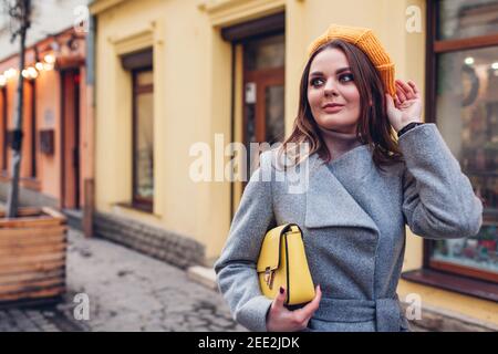 Portrait der stilvollen jungen Frau trägt gelb gestrickt Baskenmütze grauen Mantel halten Geldbörse auf der Straße im Freien. Frühling Mode weibliche Accessoires und Kleidung Stockfoto