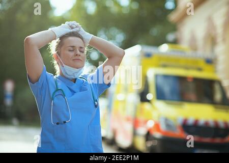 Coronavirus Pandemie. Entspannte moderne Sanitäterin Frau in Uniform mit Stethoskop und medizinische Maske Atmung im Freien in der Nähe von Krankenwagen. Stockfoto