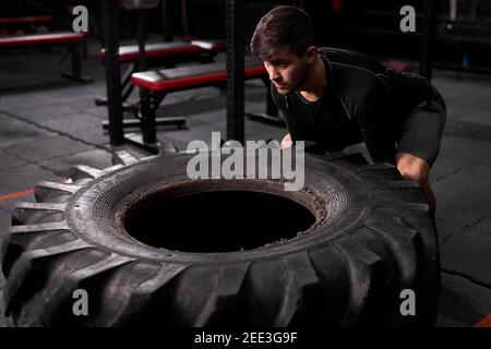 Spottman schiebt gezogenen Reifen, um Kraft im modernen Fitness-Center aufzubauen. Männlich in Sportbekleidung ist in Cross fit und Workout beschäftigt. Sport, gesunde Lebensart Stockfoto