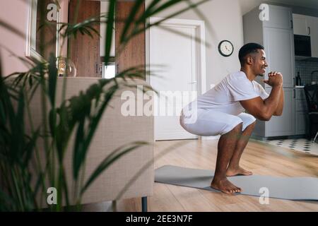 Seitenansicht des afroamerikanischen Mannes, der während des Trainings im heimischen Raum hockt und hockt. Stockfoto