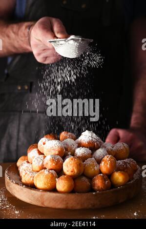 Mann streut Puderzucker auf Baked castagnole. Street Food, runde Kekse mit Zucker für den Karneval von Venedig. Traditionelle süße Backwaren während der Karnevalszeit in italien. Speicherplatz kopieren. Stockfoto