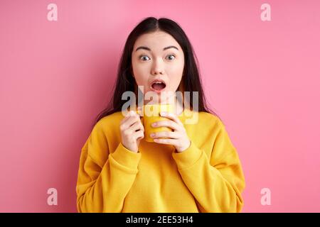 Geschockte Frau mit Tasse in den Händen, steht mit weit geöffneten Mund, überrascht von etwas. Isoliert auf rosa Hintergrund Stockfoto