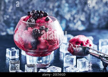 Obst- und Beereneis in einem großen schönen Glas mit Blättern aus Minze und Kugel verziert, umgeben von Eiswürfeln auf schwarzem Stein Hintergrund. Kopie, Stockfoto