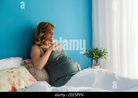 Eine schwangere Frau in einer halbsitzenden Position in ihrem Bett, während sie mit ihrem Smartphone Fotos von ihrem Bauch machte. Stockfoto