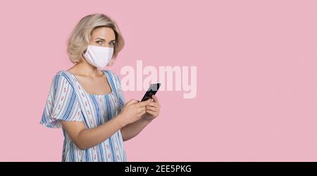 Frau mit medizinischer Maske im Gesicht und blonden Haaren ist Chatten auf Handy auf einer rosa Studio Wand mit kostenlos Platz Stockfoto