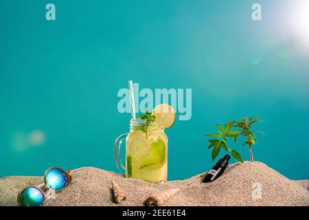 Limonade in verschiedenen Gläsern/Gläsern, CBD-Tropferöl und ein Zweig Marihuanapflanze im Sand. Sonnenbrillen und Muscheln sind von der Seite zu sehen. T Stockfoto