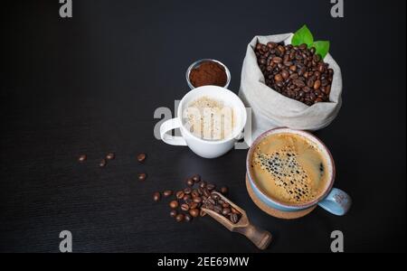 Machen Kaffee Stillleben. Kaffeetassen, geröstete Kaffeebohnen in Leinenbeutel und gemahlenes Pulver auf schwarzem Holztisch Hintergrund. Stockfoto