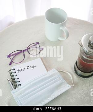 New Day - Worte auf Notizblock mit Gesichtsmaske, Kaffeepresse, Tasse und Brille im Blick. Stockfoto