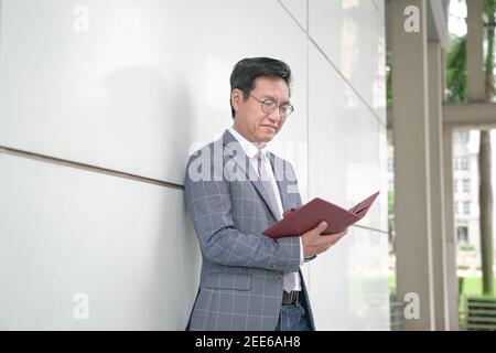 Asiatischer Geschäftsmann lehnte sich gegen whilte Wand und Blick auf sein Tagebuch. Stockfoto