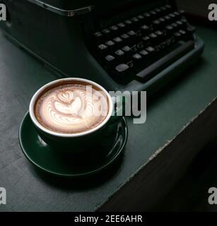 Tasse Flat White Kaffee in einer grünen Tasse neben einer alten Schreibmaschine auf einem grünen Holztisch. Nahaufnahme. Stockfoto