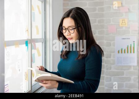 Asiatische Hochschule weiblich Student stehen und lesen Buch von der Fenster im Zimmer Stockfoto