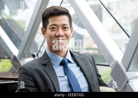 Junger lächelnder hübscher asiatisch-thailändisch-chinesischer Geschäftsmann sitzt in der Bürolounge Stockfoto