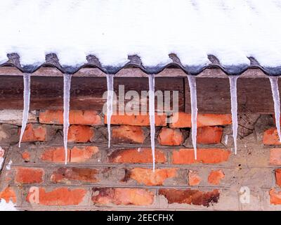 Eiszapfen aus gefrorenem Wasser auf dem Dach eines Hauses mit einer Ziegelmauer. Schnee schmilzt. Winter. Frühlingstropfen. Eiszapfen. Gefrorenes Wasser. Ziegelwand. Frostig her Stockfoto
