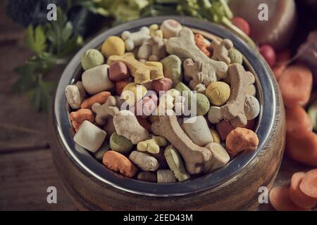 Nahaufnahme auf eine Schüssel mit gesunden ausgewogenen getrockneten Hundekekse, Kibble oder Pellets für Ihr Haustier umgeben von frischen pflanzlichen Zutaten und Knochen zu ensur Stockfoto