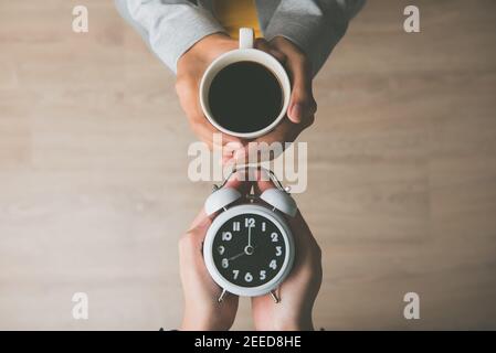 Frauen Hände halten eine Tasse Kaffee und Wecker zeigt, es ist Weckzeit am Morgen, bereit, den Tag zu beginnen Stockfoto