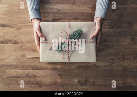 Frau, die ein weihnachtsgeschenk in braunem Papier mit rot-weißer Schnur und Kiefer, Draufsicht auf Holztisch eingewickelt Stockfoto