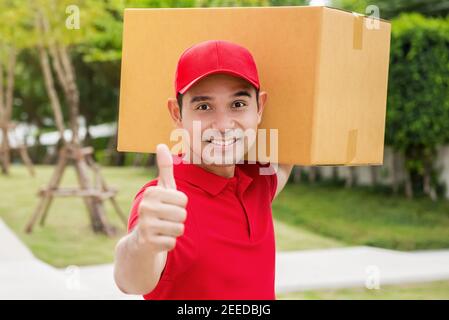 Asiatische Lieferung Mann hält ein großes Paket-Box und zeigt Daumen hoch in einem roten Poloshirt und einer roten Mütze Einheitlich Stockfoto
