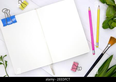 Leere Seite des Skizzenbuches mit grünen Blättern und Kunstwerkzeugen. Kreative künstlerische flache Lay mit leerer Ausbreitung von Notizbuch. Weißes Papier Skizzenbuch Mockup. Summe Stockfoto