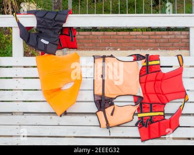 Rote und orange Rettungswesten hängen an weißen Holzzaun im Freien. Nahaufnahme Stockfoto