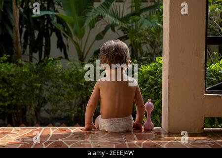 Ein charmantes Kleinkind sitzt mit einem Spielzeug auf den Stufen eines Hauses im Sonnenlicht. Rückansicht. Stockfoto