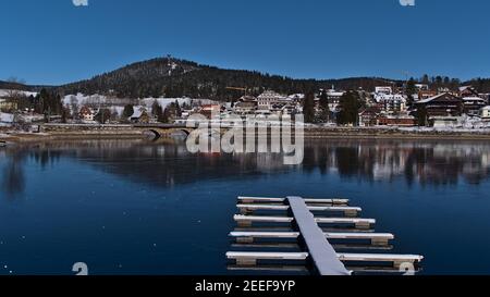 Schöner Blick auf den Kurort Schluchsee mit Spiegelungen im gefrorenen See und schneebedeckter Bootsanlegestelle an sonnigen Tagen in der Wintersaison mit blauem Himmel. Stockfoto