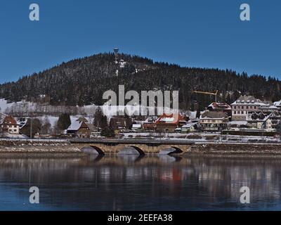 Ruhige Aussicht auf Kurort Schluchsee Dorf mit Eisenbahnbrücke auf dem gefrorenen See und Aussichtsturm Riesenbühlturm auf Hügel im Winter reflektiert. Stockfoto