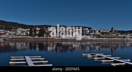 Schöner Panoramablick auf das kleine Dorf Schluchsee am Ufer des gefrorenen Sees mit schneebedeckten Landeplatten vor an sonnigen Wintertag. Stockfoto