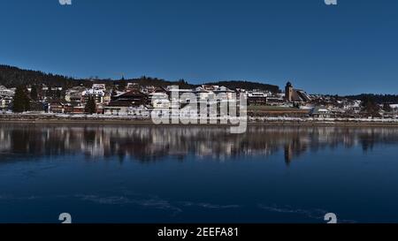 Stadtbild der Kurstadt Schluchsee mit verschneiten Gebäuden, die sich in der Wintersaison an sonnigen Tagen im gefrorenen See spiegeln. Stockfoto