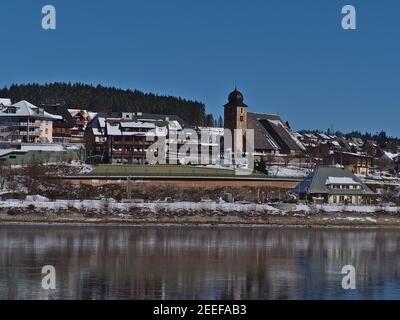 Stadtbild des Kurortes Schluchsee mit schneebedeckter Kirche St. Nikolaus, Bahnhof und Hotels spiegeln sich in der Wintersaison im gefrorenen See wider. Stockfoto