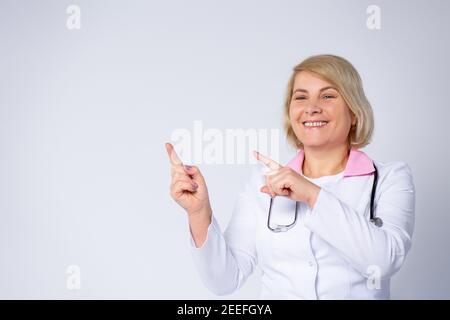 Senioren. Blonde Ärztin, lächelnd niedlich, trägt weißen Mantel und Stethoskop zur Seite zeigen mit beiden Händen auf weißem Hintergrund für Stockfoto