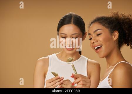 Lächelnde junge Frauen halten, Blick auf Jade Roller und Gesichts Gua Sha während zusammen posieren isoliert über beige Hintergrund. Hautpflegekonzept Stockfoto