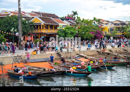 Hoi an, Vietnam - 27 Jul 2019: Historische Stadt am Flussufer mit gelben Gebäuden und Touristen. Menschen drängen sich auf dem Damm. Traditionelles Holzboot in VI Stockfoto