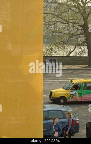 Ein hängendes gelbes Banner ergänzt ein gelbes Taxi, während zwei in Blau gekleidete Frauen an einem passenden Auto vor der Tate Britain, Millbank, London, vorbeilaufen Stockfoto